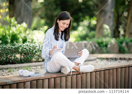 Focused young woman sitting on a wooden bench in a lush park, deeply engrossed in writing notes on a pad 113719114