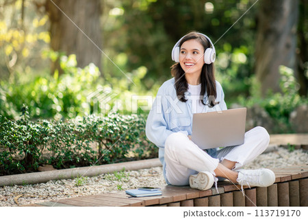 Smiling young woman sitting cross-legged on a bench in a sunlit park with headphones 113719170