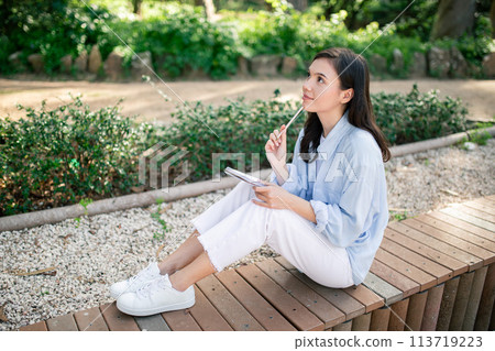Thoughtful young woman with a pencil to her lips sits on a wooden platform in a park 113719223