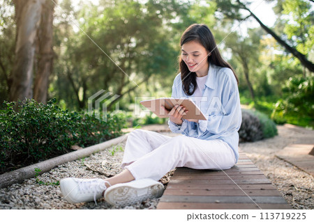 Engaged young woman reading from her notebook while sitting cross-legged on a wooden walkway Engaged young woman reading from her notebook while sitting cross-legged on a wooden walkway 113719225