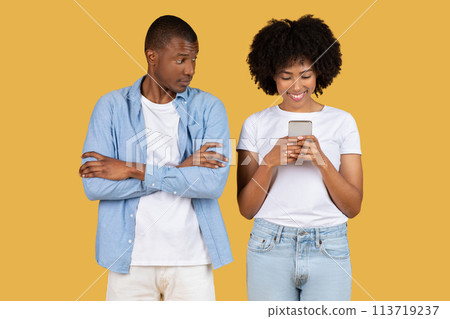 Skeptical African American man glancing at a smiling woman who is focused on her smartphone 113719237