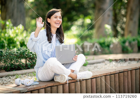 Joyful young woman waving while seated on a park bench, holding a laptop in her lap 113719260