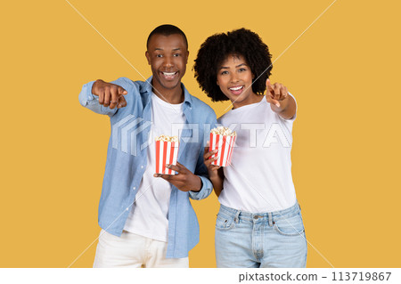 Joyful African American couple in casual clothing holding red and white striped popcorn boxes Joyful African American couple in casual clothing holding red and white striped popcorn boxes 113719867