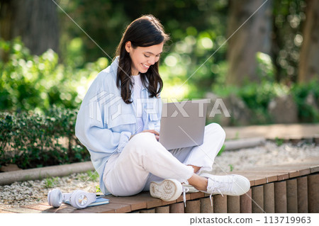 Concentrated young female sitting cross-legged on a bench in a lush garden 113719963