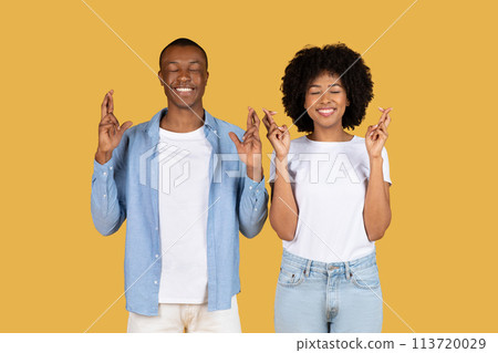 A cheerful young African American couple wearing casual clothing are crossing their fingers with eyes closed 113720029