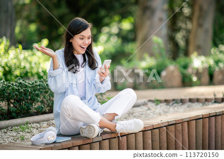 A radiant young woman sits cross-legged on a bench, animatedly reacting to her smartphone A radiant young woman sits cross-legged on a bench, animatedly reacting to her smartphone 113720150