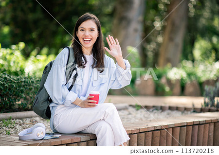 Cheerful young woman waving hello during a video call, seated on a park bench Cheerful young woman waving hello during a video call, seated on a park bench 113720218