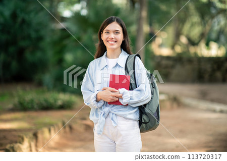 Radiant young woman in white clothes and a gray backpack holding a red book Radiant young woman in white clothes and a gray backpack holding a red book 113720317