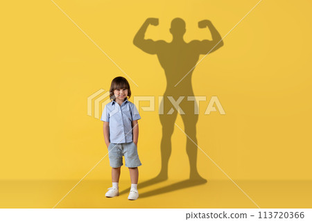 A cheerful young child stands smiling with a confident, muscular superhero shadow cast A cheerful young child stands smiling with a confident, muscular superhero shadow cast 113720366