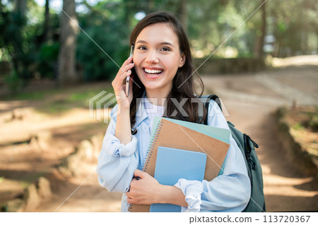 Joyful young woman engaged in a lively phone conversation while holding books Joyful young woman engaged in a lively phone conversation while holding books 113720367