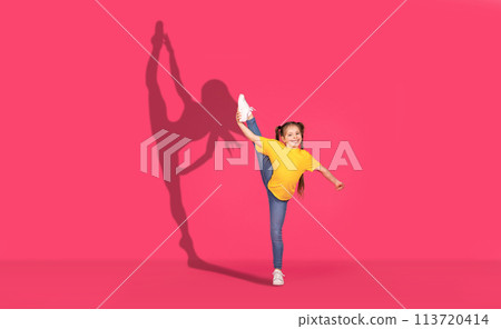 A joyful young girl in a yellow shirt and jeans performs a high leg kick with enthusiasm 113720414