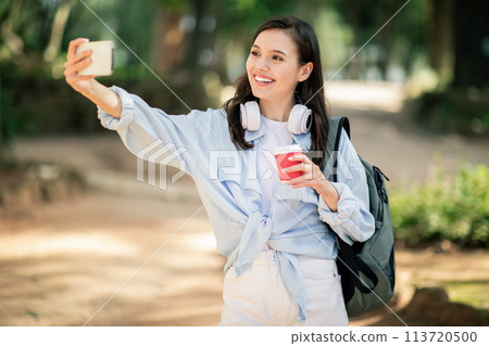 Vivacious young woman taking a selfie with her smartphone, holding a red coffee cup 113720500