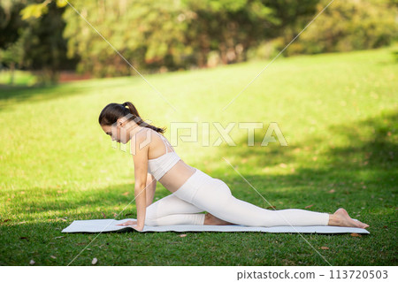Focused woman engaged in a yoga Pigeon Pose, stretching on a yoga mat in a sun-dappled park 113720503