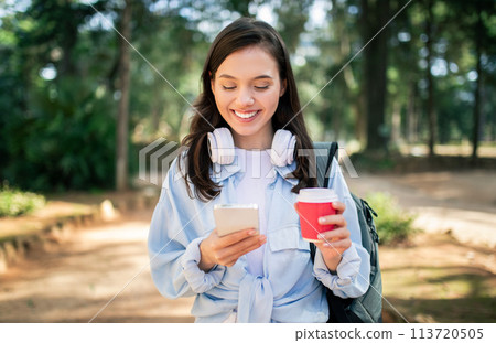 Contented young lady with headphones checking her smartphone and enjoying a warm beverage Contented young lady with headphones checking her smartphone and enjoying a warm beverage 113720505