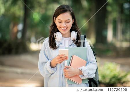Smiling young female student with headphones around her neck, looking at her smartphone 113720506
