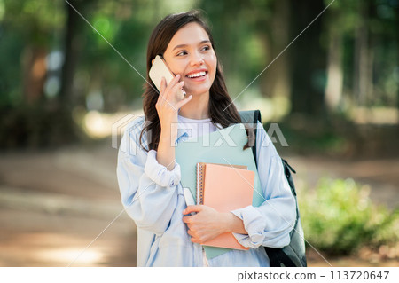 Cheerful young woman talking on a mobile phone while holding notebooks in a lush park Cheerful young woman talking on a mobile phone while holding notebooks in a lush park 113720647