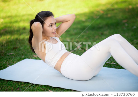 Focused woman in white activewear doing crunches on a yoga mat in a park Focused woman in white activewear doing crunches on a yoga mat in a park 113720760