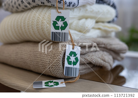 Beautifully folded woolen clothing with a recycling label lies on the table. Knitted sweaters in delicate colors with a recycling label. Eco-friendly items, biodegradable clothing, recycling. Beautifully folded woolen clothing with a recycling label lies on the table. Knitted sweaters in delicate colors with a recycling label. Eco-friendly items, biodegradable clothing, recycling. 113721392
