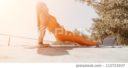 Fitness woman sea. Happy middle aged woman in orange sportswear exercises morning outdoors on yoga mat with laptop in park over ocean beach. Female fitness pilates yoga routine. Healthy lifestyle. Fitness woman sea. Happy middle aged woman in orange sportswear exercises morning outdoors on yoga mat with laptop in park over ocean beach. Female fitness pilates yoga routine. Healthy lifestyle. 113723037
