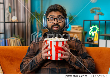 Relaxed Indian man eating popcorn watching movie enjoying film during weekend sitting on home sofa Relaxed Indian man eating popcorn watching movie enjoying film during weekend sitting on home sofa 113724925