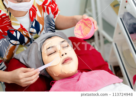 Beautiful young Asian woman getting a facial mask treatment at the beauty salon. Facial skincare. 113724948