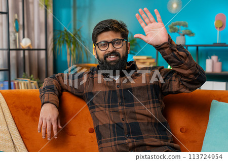 Indian man smiling friendly at camera, waving hands gesturing hello, hi, greeting at home on sofa 113724954