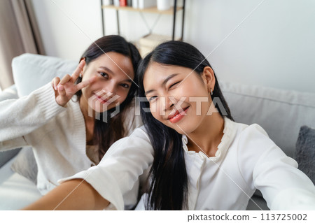 Two women are smiling and taking a selfie sitting on the sofa in the living room at home. 113725002