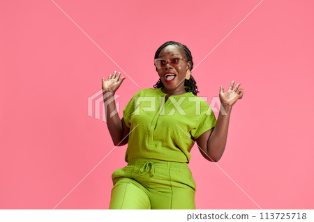 Existed young African-American woman in casual attire raising hands in delight against pastel pink studio background. Existed young African-American woman in casual attire raising hands in delight against pastel pink studio background. 113725718