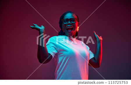 Existed young African-American woman in casual attire raising hands in delight in red neon light against gradient studio background. Existed young African-American woman in casual attire raising hands in delight in red neon light against gradient studio background. 113725753