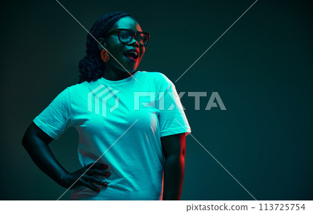 Emotional young African-American woman hold hand on hip and looking away in red neon light against gradient studio background. Emotional young African-American woman hold hand on hip and looking away in red neon light against gradient studio background. 113725754