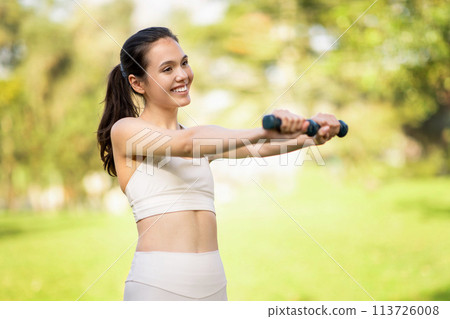 Radiant young woman with a beaming smile exercises with hand weights in a lush green park 113726008