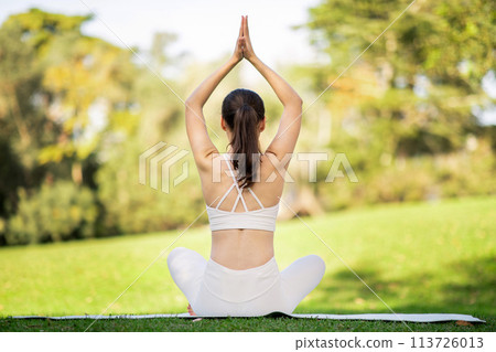 Viewed from behind, a woman in a seated yoga pose extends her arms gracefully overhead 113726013