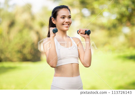 Cheerful young woman with a bright smile lifting dumbbells, enjoying her fitness routine in the fresh air 113726143