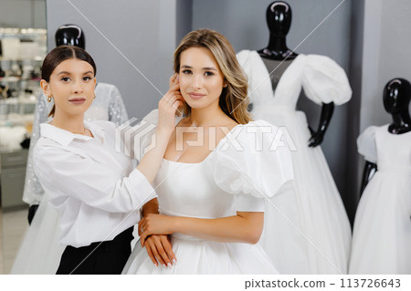 The bride-to-be came to a wedding dress salon to buy a dress. A beautiful woman chooses a wedding dress in the salon, and an assistant helps the bride put on an earring The bride-to-be came to a wedding dress salon to buy a dress. A beautiful woman chooses a wedding dress in the salon, and an assistant helps the bride put on an earring 113726643