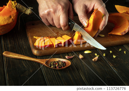 Chef hands use a knife to cut pumpkin into small pieces on a cutting board for baking a pie. American national dish cooking concept Chef hands use a knife to cut pumpkin into small pieces on a cutting board for baking a pie. American national dish cooking concept 113726776