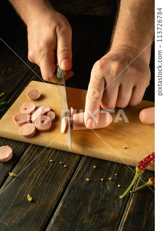 The chef cuts the sausage into small pieces with a knife. Preparing a delicious breakfast with milk sausage and spices on the kitchen table in a public house The chef cuts the sausage into small pieces with a knife. Preparing a delicious breakfast with milk sausage and spices on the kitchen table in a public house 113726784