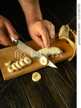 A cook uses a knife to slice a ripe banana to prepare a fruit dish on the kitchen table. Place for advertising on a dark background 113726785