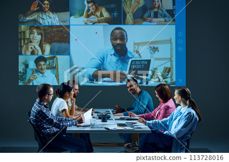Workers sitting in office boardroom participate in virtual meeting, with remote colleagues via online video call 113728016