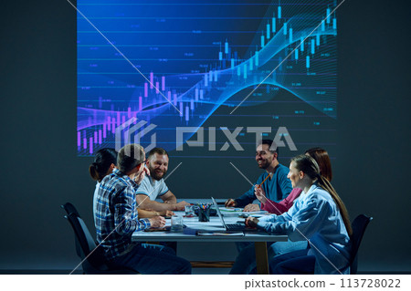 Teammates in a corporate environment sit around a conference table, focusing on a digital stock chart displayed on a monitor in the boardroom. 113728022