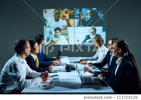 Employees sit in modern office boardroom, discussing projects and documents over a video call with colleagues and trainees 113728126