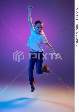 Summer break. Young boy in casual outfit jumping raising hands of joy and fun in neon light against purple gradient background. Summer break. Young boy in casual outfit jumping raising hands of joy and fun in neon light against purple gradient background. 113729728