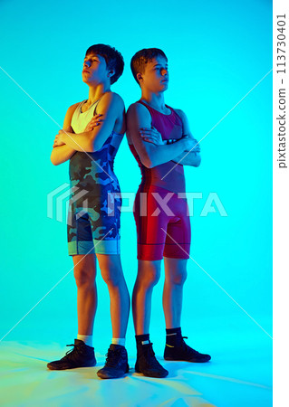 Two male wrestlers standing, face off in physical contest, each aiming to win the other against blue background in neon light Two male wrestlers standing, face off in physical contest, each aiming to win the other against blue background in neon light 113730401