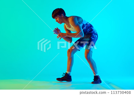 Athletic young man in blue uniform standing in wrestling pose, ready to fight against blue background in neon light 113730408