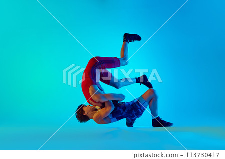 Young men, wrestlers compete in a high-energy match, demonstrating skill and strategy against blue background in neon light 113730417