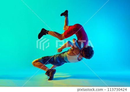 Male wrestlers engage in physical battle, each striving for victory, competing, training against blue background in neon light 113730424