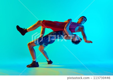 Young men, athletes demonstrate focus and strength as they engage in competitive wrestling match against blue background in neon light 113730446