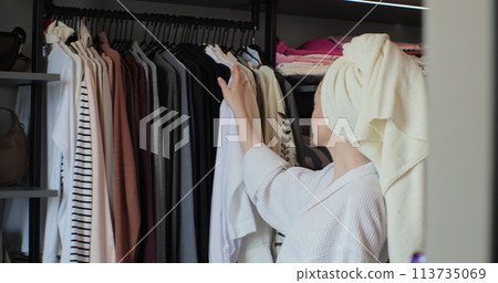 Young attractive woman wearing a bathrobe and a towel choosing her outfit clothes in closet in bedroom at home. Young attractive woman wearing a bathrobe and a towel choosing her outfit clothes in closet in bedroom at home. 113735069