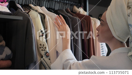 Woman wearing a bathrobe and a towel on her head choosing clothes from rack with hangers inside room. Lady looks to clothes, decides what to wear. Lifestyle women relax at home concept. Woman wearing a bathrobe and a towel on her head choosing clothes from rack with hangers inside room. Lady looks to clothes, decides what to wear. Lifestyle women relax at home concept. 113735071