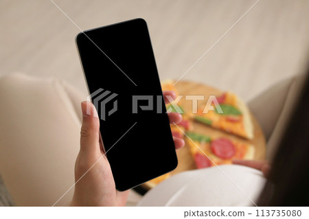 Unrecognizable pregnant female eating pizza and using mobile phone holding smartphone with black blank screen advertisement of delivery service of fast food 113735080