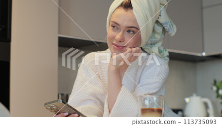 Portrait of happy woman wearing a bathrobe and a towel on her head using smartphone in the kitchen sending message on social media sharing. The morning is routine. Portrait of happy woman wearing a bathrobe and a towel on her head using smartphone in the kitchen sending message on social media sharing. The morning is routine. 113735093